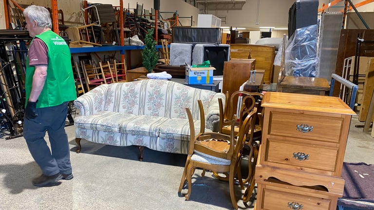 Person in green shirt examining furniture at a warehouse sale including a wrapped sofa, wooden chairs, and dresser
